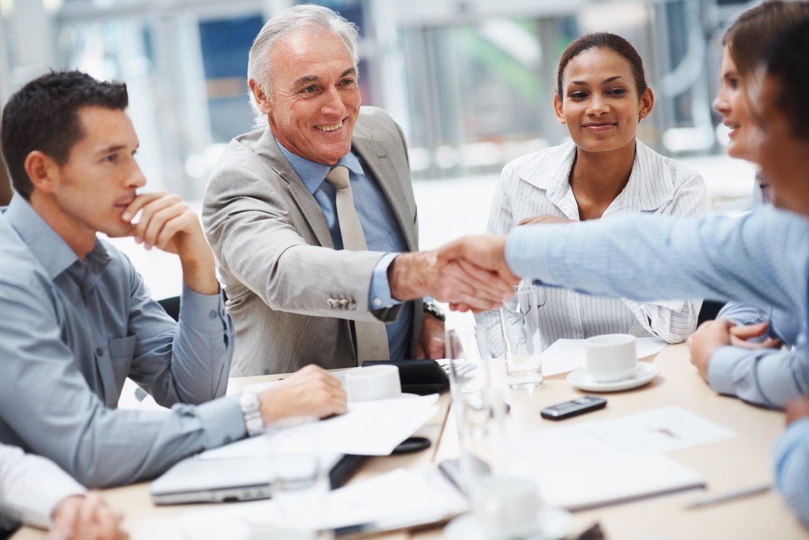 Diverse workplace collaboration, five individuals from various departments, two shaking hands across the table.